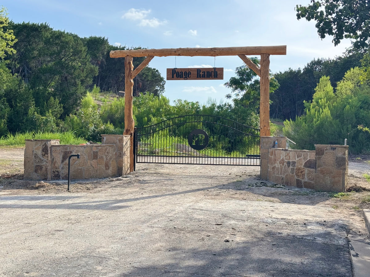 Custom arched steel ranch entry gate with plasma-cut brand, cedar log header and stone columns — built and installed by Prestigious Metal Works, Waco TX